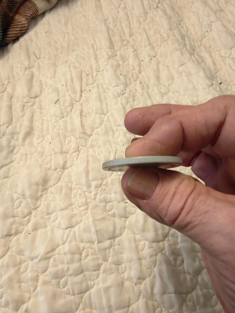 May include: A silver coin held between fingers, showing the edge of the coin. The coin has a reeded edge and appears to be a quarter. The background is a cream-colored, quilted fabric.