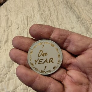 May include: A white and gold commemorative coin with the text "One Year" and the number "1". The coin has a decorative gold border with ornate swirls. The coin is held in a hand.