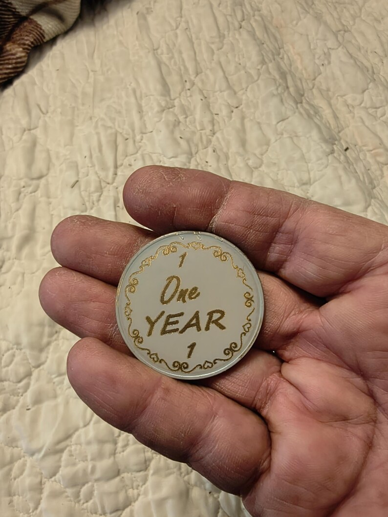 May include: A white and gold commemorative coin with the text "One Year" and the number "1". The coin has a decorative gold border with ornate swirls. The coin is held in a hand.