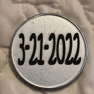 以下が含まれることがあります： 丸い銀色のオブジェクトで、太字の黒いフォントで「3-21-2022」の日付が印刷されています。 オブジェクトには金属製の境界線があり、わずかに傷が付いているように見えます。 背景はキルトのオフホワイトの生地です。