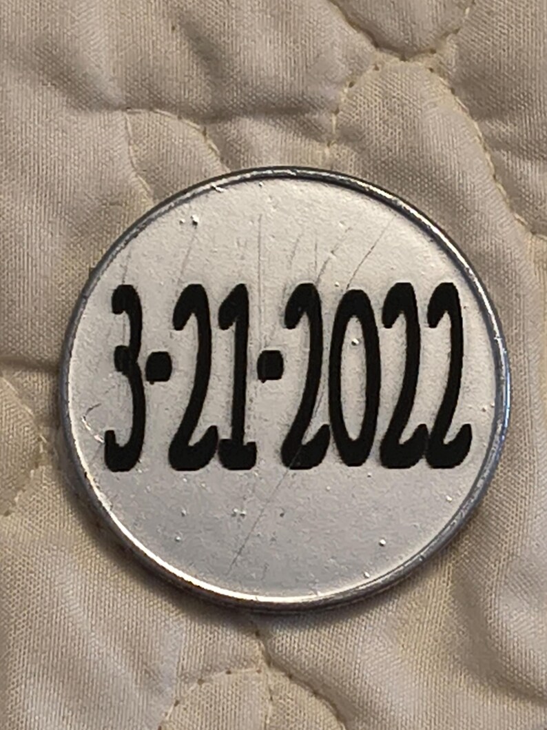 以下が含まれることがあります： 丸い銀色のオブジェクトで、太字の黒いフォントで「3-21-2022」の日付が印刷されています。 オブジェクトには金属製の境界線があり、わずかに傷が付いているように見えます。 背景はキルトのオフホワイトの生地です。