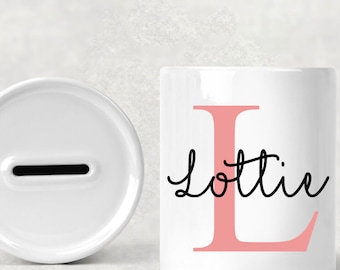 May include: White ceramic piggy bank with a slot for coins. The bank features a large pink letter "L" and the name "Lottie" in black cursive script. The design is simple and elegant.