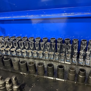 May include: A collection of chrome and black metal socket wrench attachments organized on black plastic rails inside a bright blue metal tool chest. The set includes various sizes of cylindrical sockets with hexagonal openings for mechanical work.