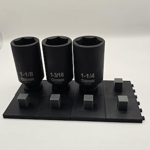 May include: Three black Quinn impact sockets in sizes 1-1/8, 1-3/16, and 1-1/4 inches, labeled CR-MO. The sockets are arranged upright on a black modular plastic organizer tray featuring square pegs and a textured surface finish.