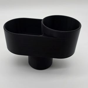 May include: A matte black plastic cup holder insert featuring a dual-compartment design. The unit consists of a circular cup holder section attached to an elongated oval storage tray, supported by a single cylindrical base underneath the structure.