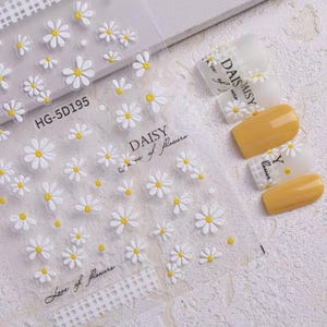 Könnte beinhalten: Transparente Folie mit Daisy-Nagelkunst-Aufklebern mit weißen und gelben Blumenmotiven. Die Folie enthält den Text "DAISY Love of flowers" und den Code "HG-5D195". Mehrere Nägel sind mit gelbem Lack und Daisy-Designs bemalt.