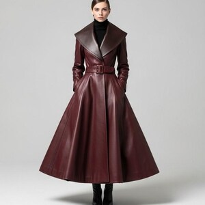May include: A full-length, burgundy leather coat with a wide collar and belted waist. The coat features a flared skirt and long sleeves. The model is wearing a black turtleneck and black boots. The coat has a sophisticated, tailored design.