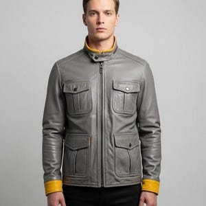 Op de afbeelding: Een grijze leren jas met een gele kraag en manchetten. De jas heeft vier voorzakken en een ritssluiting. Het model draagt een zwarte broek.