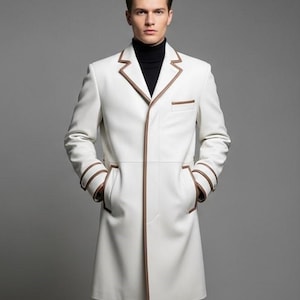 Puede incluir: Un abrigo blanco con ribetes y detalles marrones. El abrigo presenta un cuello con solapas, un diseño de un solo pecho y bolsillos. Las mangas tienen ribetes marrones alrededor de los puños. La persona lleva un cuello alto negro.