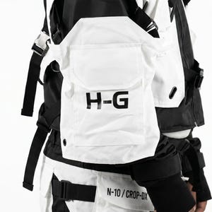Pu&ograve; includere: Gilet tattico bianco e nero con pi&ugrave; tasche e cinghie. Il gilet presenta le lettere "H-G" stampate in nero su una tasca frontale. Il design include una combinazione di pannelli bianchi e neri, con cinghie e fibbie regolabili.