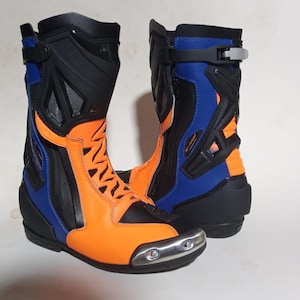 Peut inclure: Une paire de bottes de moto mi-mollet présentant un coloris orange vif, bleu roi et noir. Les bottes comprennent un embout métallique argenté, des panneaux de protection noirs moulés et un système de fermeture à boucle sur le dessus.