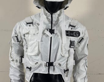 Op de afbeelding: Witte cropped jas met een capuchon en meerdere zakken, met zwarte gespen en riemen. De jas heeft de tekst "H-G" op de borst en andere tekst op de mouwen. De jas wordt getoond op een zwarte mannequin.