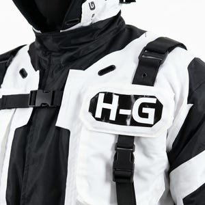 Pu&ograve; includere: Giacca bianca e nera con cappuccio. La giacca presenta le lettere "H-G" sul petto e cinghie nere con fibbie. Il design &egrave; un mix di pannelli bianchi e neri.