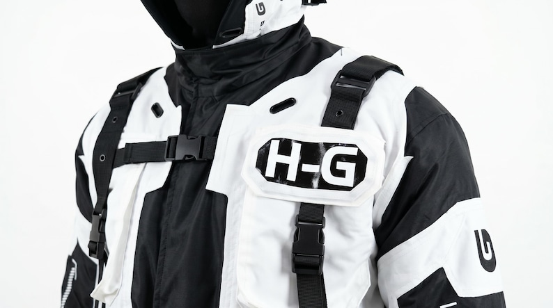 Pu&ograve; includere: Giacca bianca e nera con cappuccio. La giacca presenta le lettere "H-G" sul petto e cinghie nere con fibbie. Il design &egrave; un mix di pannelli bianchi e neri.