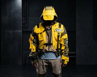 Può includere: Giacca con cappuccio gialla e verde oliva con più tasche e cinghie nere. La giacca ha il testo "H-G" sul petto. La persona indossa una camicia azzurra e pantaloni marroni. Lo sfondo è un ambiente industriale scuro.