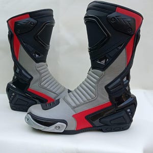 Puede incluir: Un par de botas de moto en negro, gris y rojo. Las botas presentan un diseño de caña alta con placas de plástico negro reforzadas en espinillas, talones y tobillos, paneles de cuero gris y detalles rojos en los laterales y bordes superiores.