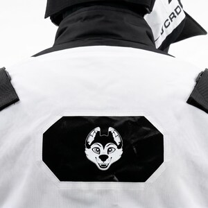 Pu&ograve; includere: Giacca bianca e nera con logo a forma di muso di husky bianco e nero sul retro. La giacca presenta dettagli neri e cinturini neri sulle spalle. Il colletto &egrave; nero e il capo sembra essere realizzato in un materiale resistente.