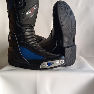 Peut inclure: Une paire de bottes de moto noires avec des panneaux latéraux bleus et des curseurs d'orteils argentés. Les bottes présentent le nom de marque REXEL en blanc sur la tige, une construction en cuir noir et une semelle en caoutchouc texturé.