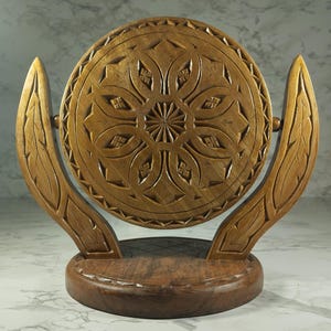 May include: A decorative wooden piece with a circular, carved design. The piece has a round base and two curved supports. The carved design features floral and geometric patterns. The wood has a warm, natural tone, and the piece is likely handmade.