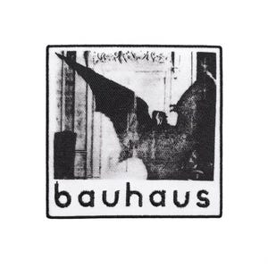 Puede incluir: Un parche en blanco y negro con una imagen estilizada de una figura parecida a un pájaro o un murciélago. La palabra "bauhaus" está impresa en blanco en la parte inferior. El parche tiene un borde blanco.