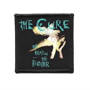 Puede incluir: Parche cuadrado negro con el nombre de la banda "The Cure" en turquesa y el título del álbum "The Head on the Door". El diseño incluye una imagen surrealista en amarillo y blanco. El parche tiene un borde cosido.