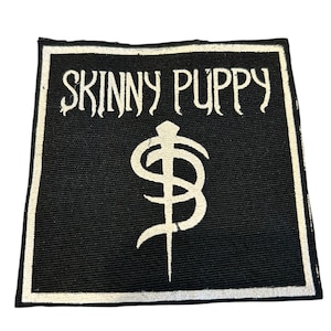 Peut inclure: Écusson carré noir avec une bordure blanche. L'écusson porte l'inscription "SKINNY PUPPY" en blanc, au-dessus d'un symbole stylisé blanc. L'écusson est probablement un accessoire pour vêtements ou sacs.