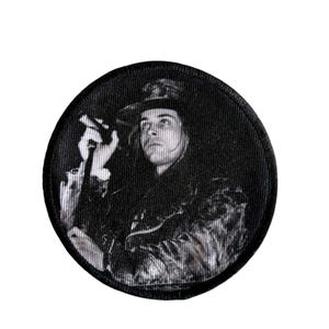 May include: Black and white circular patch featuring a vintage image of a person wearing a hat and holding a microphone. The patch has a textured appearance and a black border. The image is a grayscale photograph.