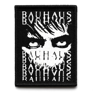 Puede incluir: Parche rectangular negro con texto blanco y un rostro estilizado. La palabra "BAUHAUS" está impresa verticalmente en los lados y horizontalmente en la parte superior e inferior. El rostro tiene ojos grandes.