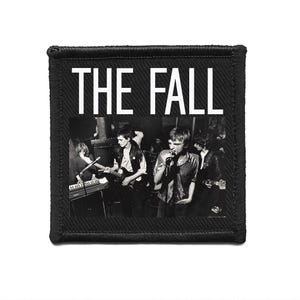 May include: Black and white patch with the text "THE FALL" in white. The image shows a band performing live, with a singer and musicians playing instruments. The patch has a black border.