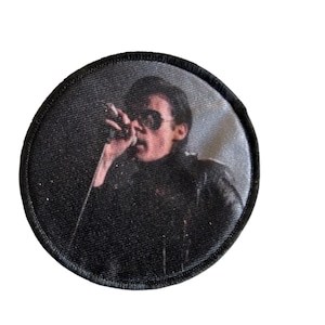 Può includere: Toppa rotonda nera con un'immagine in bianco e nero di una persona con occhiali da sole e giacca di pelle nera, che tiene un microfono. La toppa ha un bordo nero ed è probabilmente una toppa da stirare o da cucire.