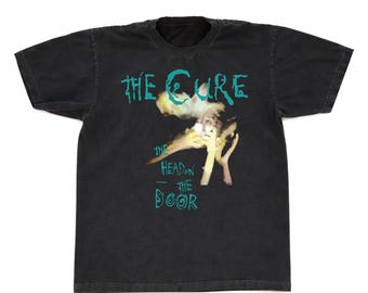 May include: Black t-shirt featuring the band name "The Cure" and album title "The Head on the Door" in teal lettering. The graphic includes a surreal image of a face and hands. The shirt has a vintage, worn look.
