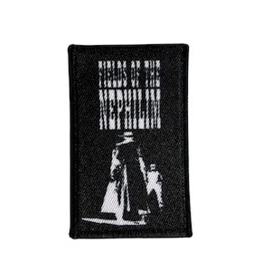May include: Black rectangular patch featuring white text and a graphic. The text reads "FIELDS OF THE NEPHILIM". The graphic shows two figures. The patch is designed for application on clothing or accessories.