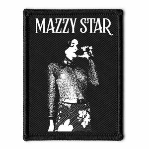 May include: Black rectangular patch with the text "MAZZY STAR" in white, above a white illustration of a person holding a microphone. The person is wearing a patterned top and skirt. The patch has a black border.