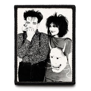 May include: Black and white patch featuring a portrait of two people. One person is wearing a patterned shirt and suspenders, the other a t-shirt with a graphic of a horned mask. The patch has a black border.