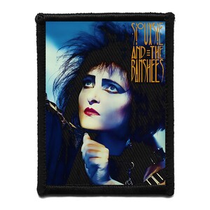 Puede incluir: Parche bordado negro con un retrato de Siouxsie Sioux, con maquillaje llamativo y cabello oscuro. El parche muestra el texto "Siouxsie and the Banshees" en dorado sobre un fondo azul. El parche tiene un borde negro.