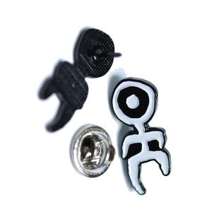 Puede incluir: Dos pines de esmalte con un diseño abstracto en blanco y negro. Un pin es negro con un pasador, y el otro es blanco con contornos negros. Los pines tienen un estilo único y minimalista.