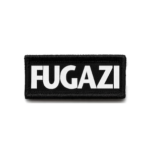 May include: Black rectangular patch with the word "FUGAZI" in bold white capital letters. The patch has a black border with a stitched texture. The patch is centered on a white background.