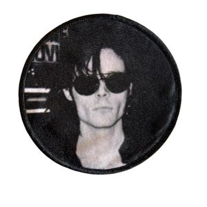 May include: Circular black and white patch with a portrait of a person wearing sunglasses. The patch has a black border and a textured finish. The image is a close-up of the person's face.