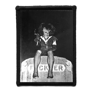 May include: Black and white patch depicting a person in a dark outfit, fishnet stockings, and a cross necklace, seated on a tombstone. The tombstone has the word "F*CKER" on it. The patch has a black border.