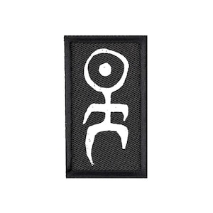 Może przedstawiać: Prostokątna czarna naszywka z białym abstrakcyjnym symbolem. Symbol przypomina ludzika z dużą okrągłą głową i zakrzywionymi ramionami i nogami. Naszywka ma czarną obwódkę.
