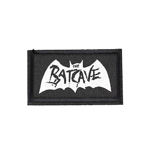 May include: A black rectangular patch featuring a white bat symbol and the words "THE BATCAVE". The text is in a distressed style. The patch has a black border and is probably made of fabric.