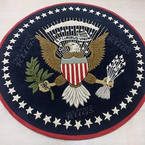 Puede incluir: Alfombra redonda con el emblema del águila americana. La alfombra tiene un fondo azul marino y un borde rojo. El emblema incluye un águila con las alas extendidas, sosteniendo una rama de olivo y flechas. El texto "E PLURIBUS UNUM" es visible.
