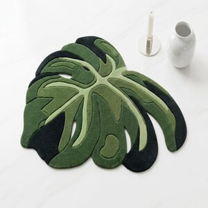 Puede incluir: Una alfombra tufting con forma de hoja de monstera grande, que presenta un diseño en capas con varios tonos de verde oscuro, verde oliva y negro. La alfombra tiene una superficie texturizada con surcos profundos que definen las nervaduras.