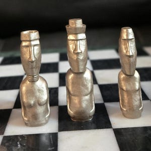 May include: A unique chess set featuring gold-colored pieces shaped like Moai statues. The pieces are arranged on a black and white checkered marble board. The background is a blurred dark surface.