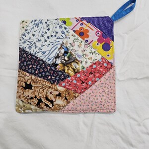 May include: A patchwork potholder with a variety of fabric patterns, including floral, cat, and geometric designs. The potholder has a blue loop for hanging and is approximately 8 inches square.