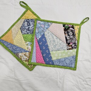 May include: Two handmade, square pot holders with a patchwork design. Each pot holder features a variety of fabric patterns in shades of blue, pink, yellow, and green, with a green border and a loop for hanging.