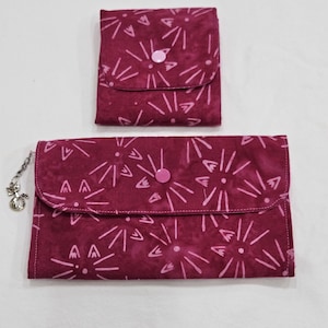 May include: Two-piece set of burgundy fabric pouches with a floral pattern. The larger pouch has a flap closure with a pink button and a silver cat charm. The smaller pouch also has a flap closure with a pink button.