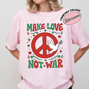 Könnte beinhalten: Hellrosa T-Shirt mit dem Schriftzug "MAKE LOVE NOT WAR" in Grün und Rot. Das Design beinhaltet ein rotes Friedenszeichen mit einer weißen Taube, die einen Olivenzweig trägt. Das T-Shirt ist von der Marke Comfort Colors.