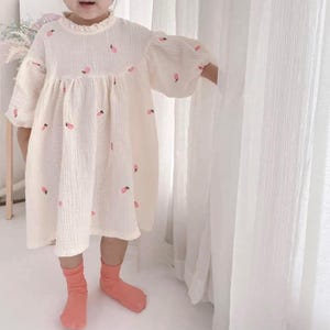 Puede incluir: Un vestido color crema con bordados de fresas rosas. El vestido presenta un cuello con volantes, mangas largas con puños abullonados y una falda fruncida. La niña lleva calcetines color coral.