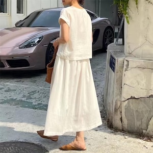 May include: A white linen two-piece outfit, comprising a short-sleeved top and a long skirt. The outfit is worn with brown woven sandals and a woven tote bag. A car is parked in the background.